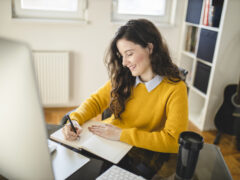 A woman with brown curls and a yellow sweater is working from home and taking notes