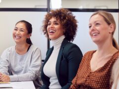 Three women from different backgrounds sit in an office and laugh.