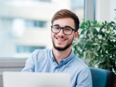 A young man in glasses is working on his laptop while smiling.